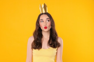 Beautiful young woman with princess crown sending air kiss on yellow background