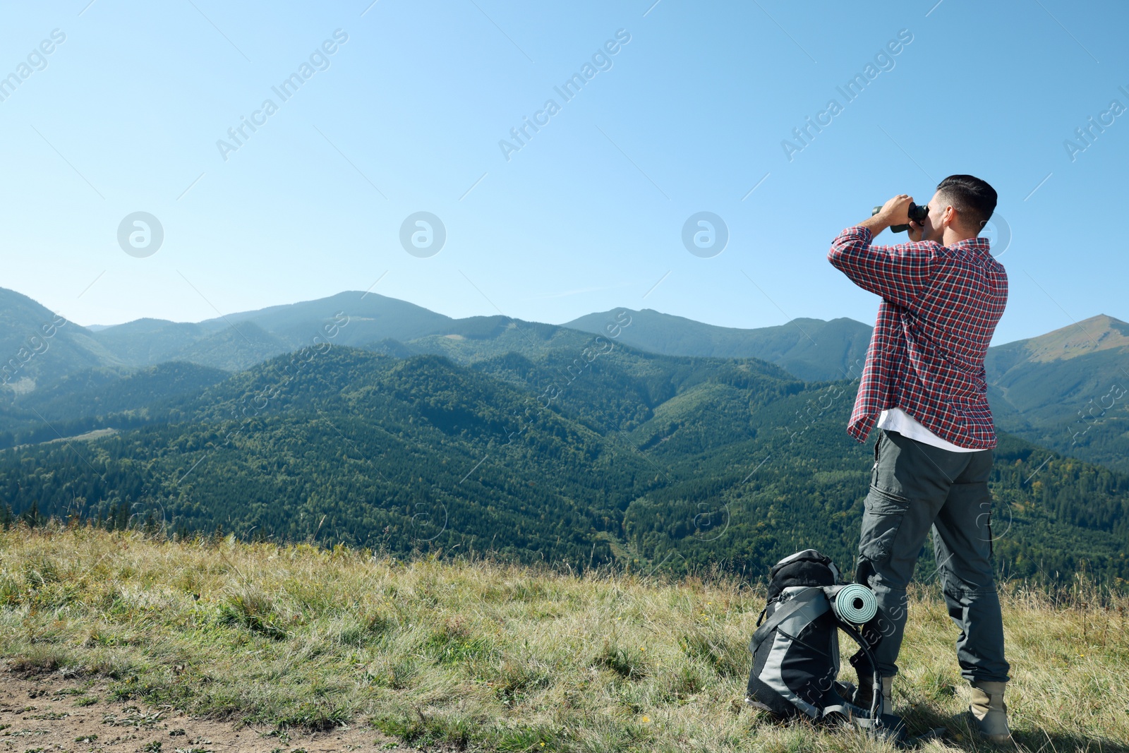 Photo of Tourist with hiking equipment looking through binoculars in mountains, back view