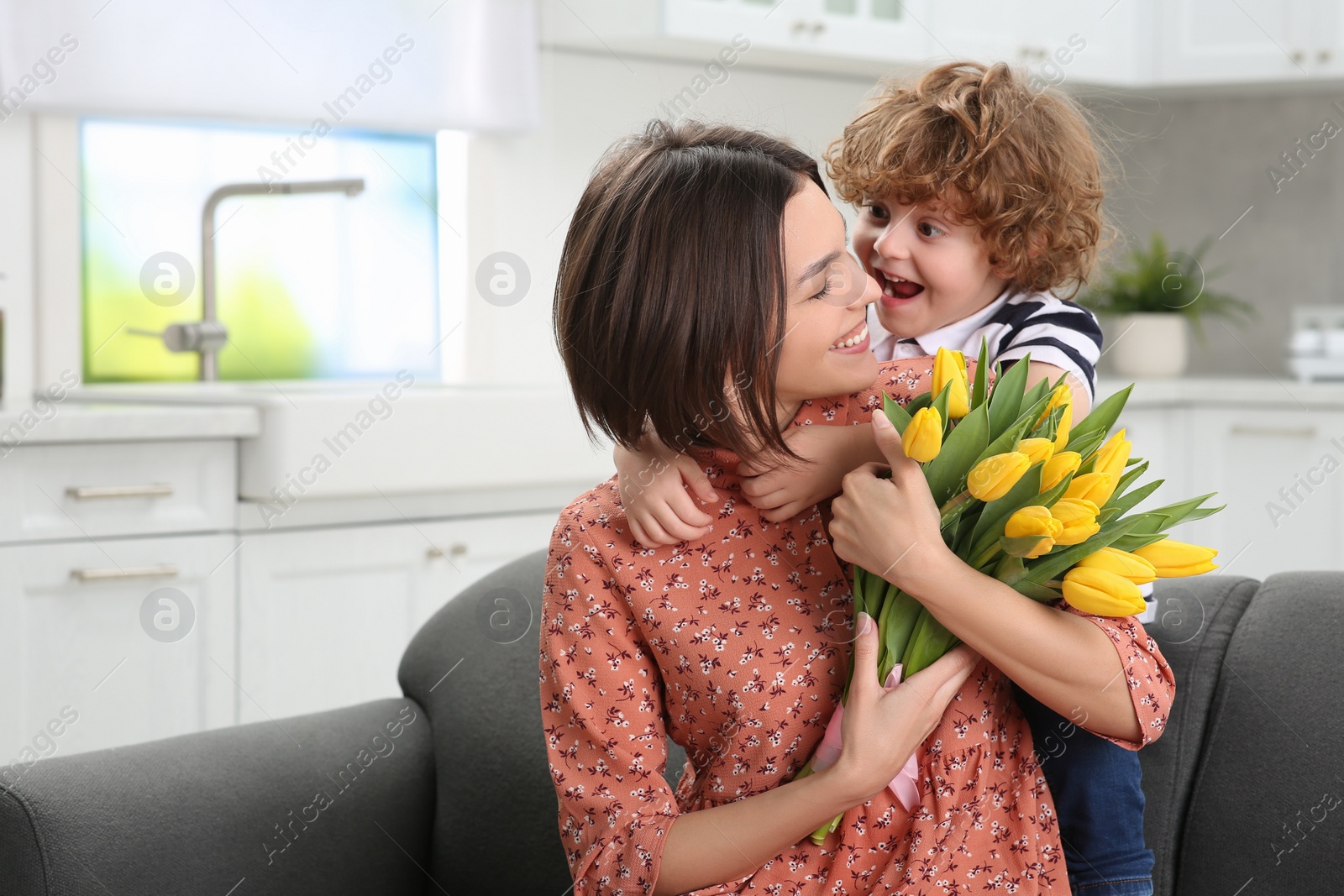 Photo of Little son congratulating his mom with Mother`s day at home. Woman holding bouquet of yellow tulips