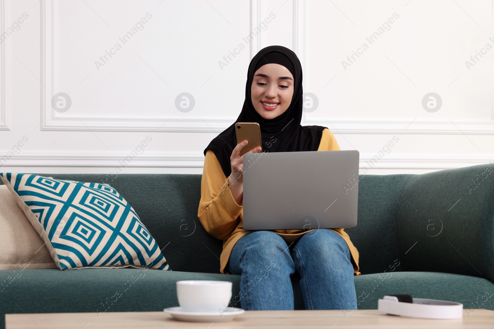 Photo of Muslim woman in hijab using laptop and smartphone on sofa indoors