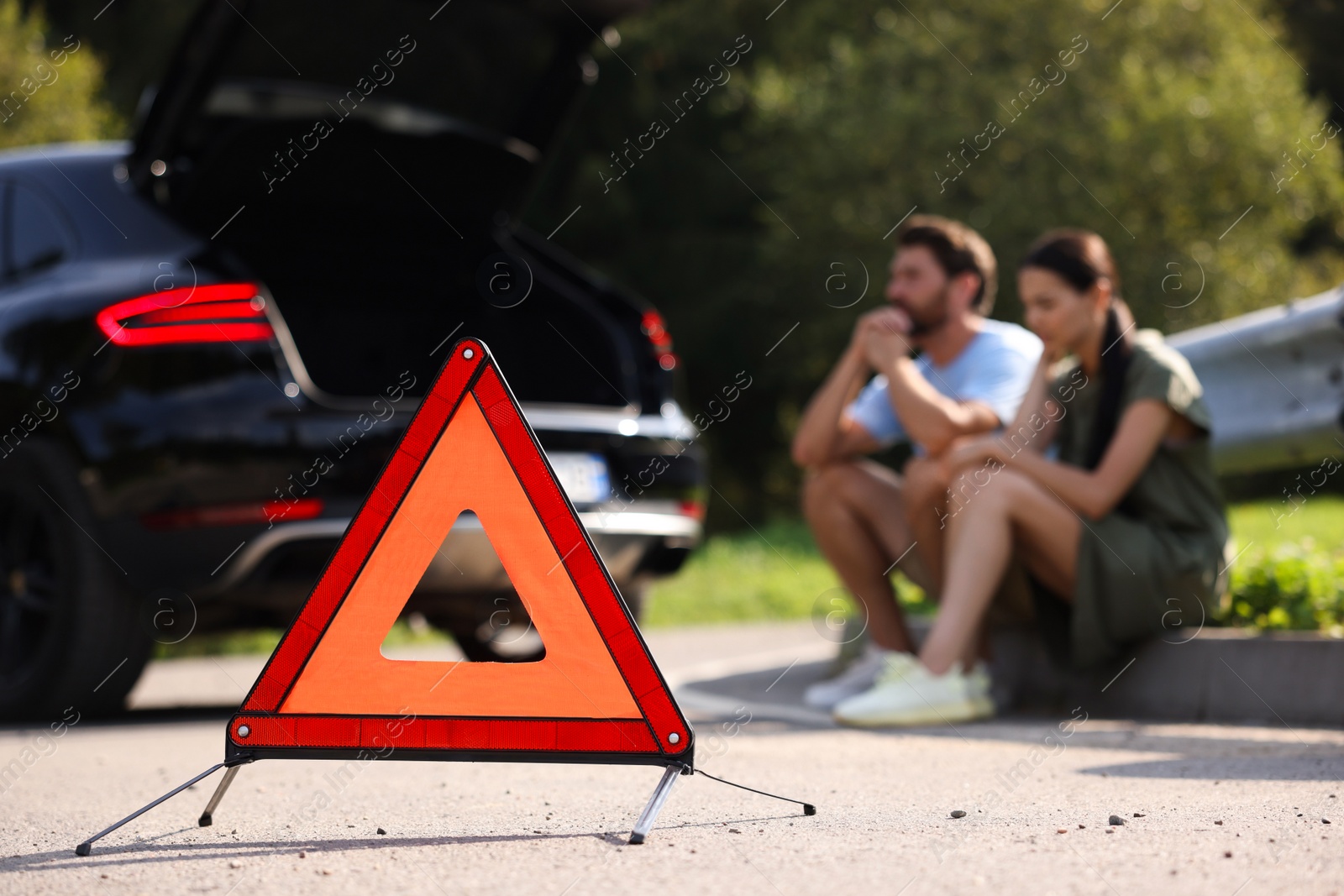 Photo of Couple sitting near broken car on roadside outdoors, focus on warning triangle