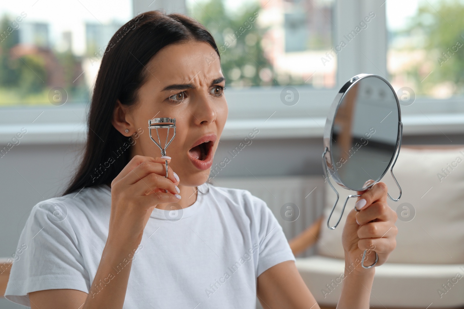 Photo of Emotional young woman with mirror using eyelash curler indoors