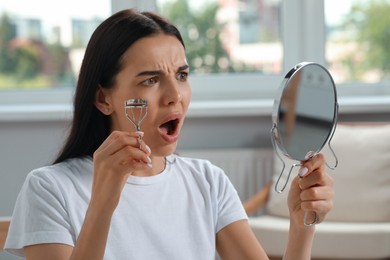 Emotional young woman with mirror using eyelash curler indoors