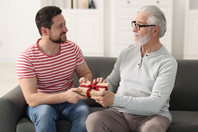 Photo of Son giving gift box to his dad on sofa at home