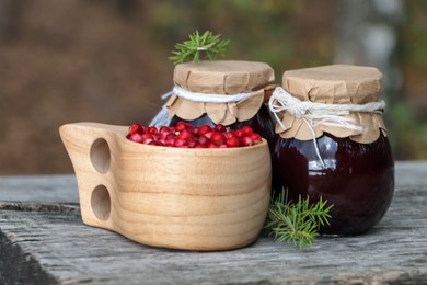 Photo of Tasty lingonberry jam in jars and cup with red berries on wooden table outdoors