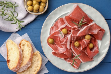 Photo of Slices of tasty cured ham, rosemary, bread and olives on blue wooden table, flat lay