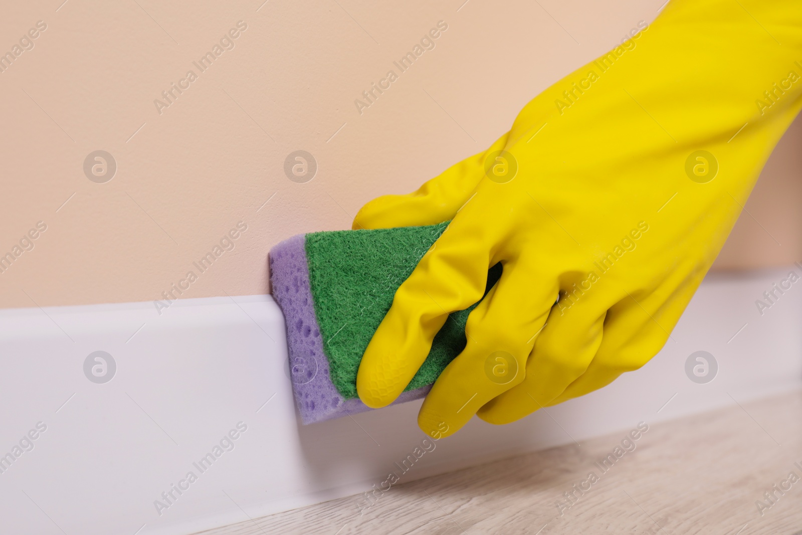 Photo of Woman in protective glove cleaning plinth with sponge indoors, closeup