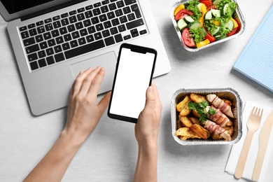 Photo of Top view of woman holding smartphone over white wooden table with lunchboxes,  mockup for design. Healthy food delivery