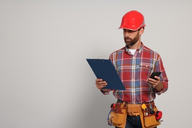 Photo of Professional builder in hard hat with clipboard, phone and tool belt on light background, space for text
