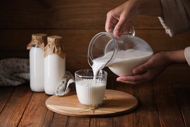 Photo of Woman pouring fresh milk from jug into glass at wooden table, closeup