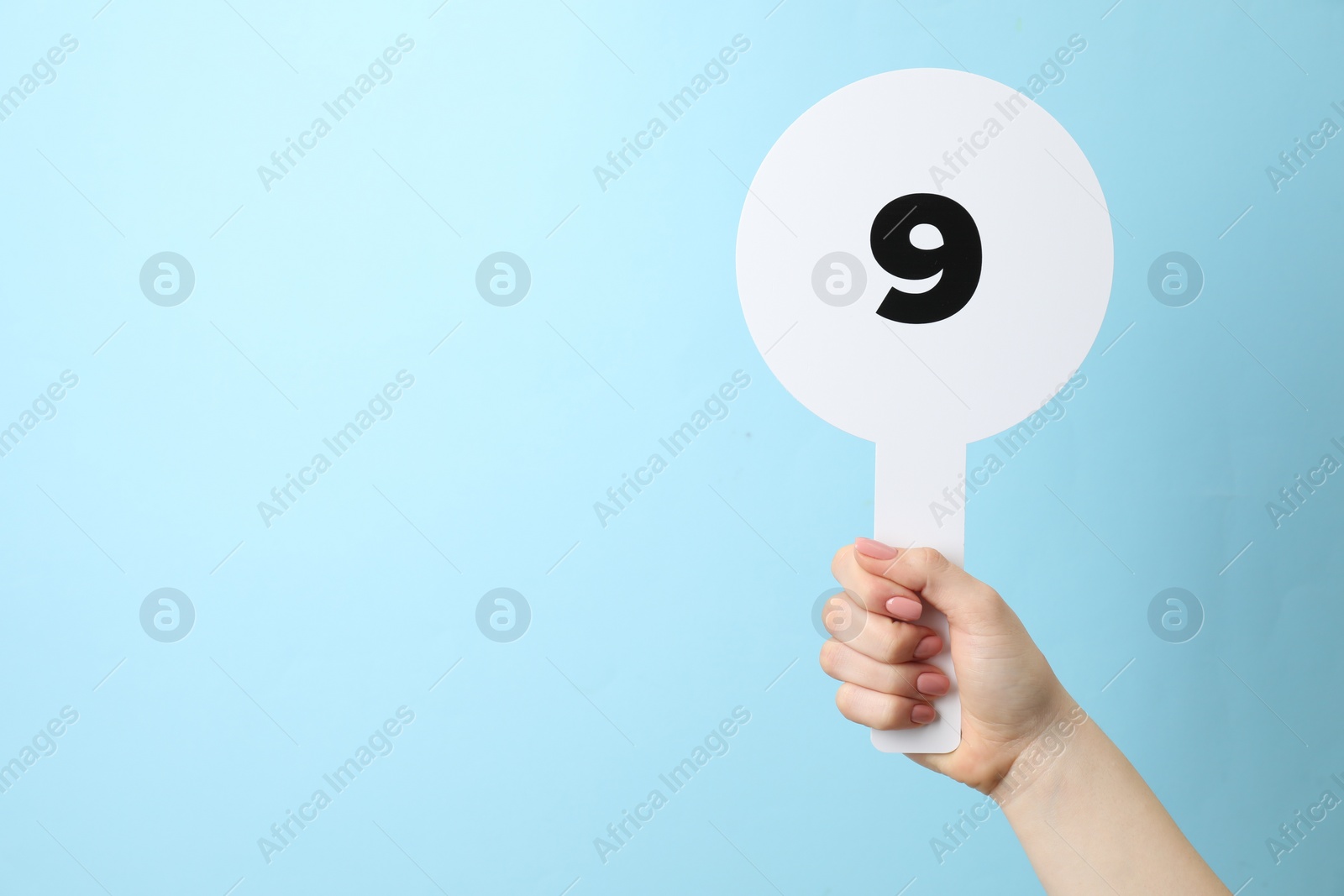 Photo of Woman holding auction paddle with number 9 on light blue background, closeup. Space for text