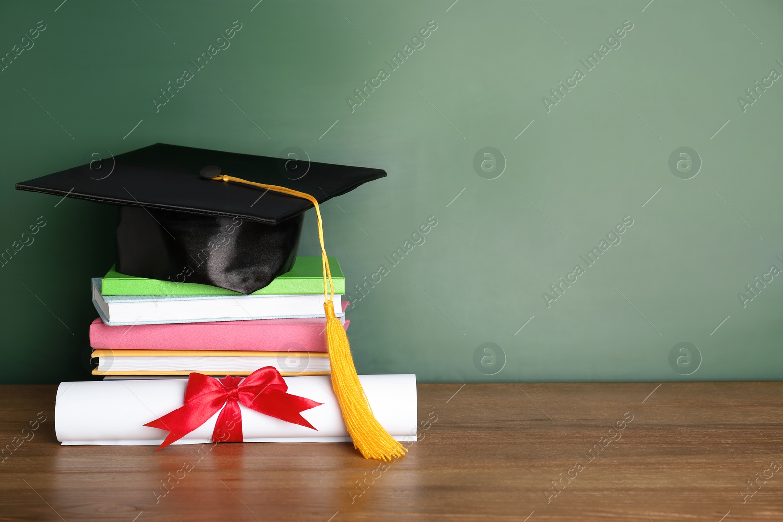 Photo of Graduation hat with books and diploma on table near chalkboard. Space for text