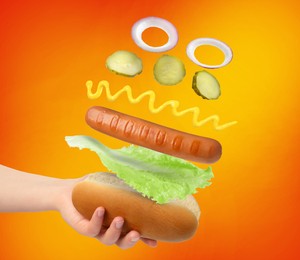 Image of Woman making hot dog on orange gradient background, closeup. Ingredients levitating over bun
