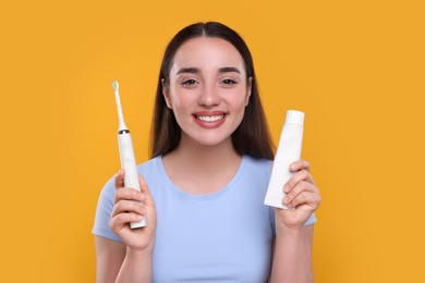 Happy young woman holding electric toothbrush and tube of toothpaste on yellow background