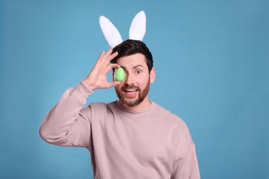 Happy man in cute bunny ears headband covering eye with Easter egg on light blue background