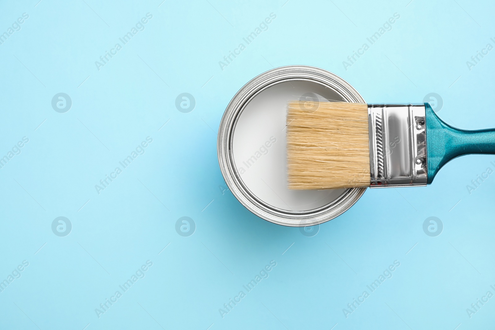 Photo of Open can with white paint and brush on blue background, top view. Space for text