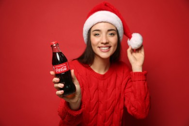 MYKOLAIV, UKRAINE - JANUARY 27, 2021: Young woman in Christmas hat holding bottle of Coca-Cola against red background, focus on hand