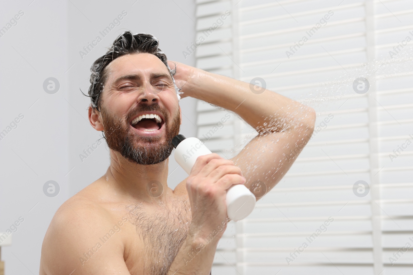 Photo of Happy man with bottle of shampoo singing in shower. Space for text