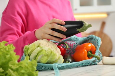 Photo of Woman taking eggplant out from string bag at light marble table, closeup