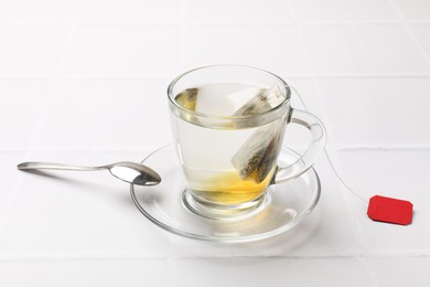 Tea bag in cup with hot drink and spoon on white tiled table