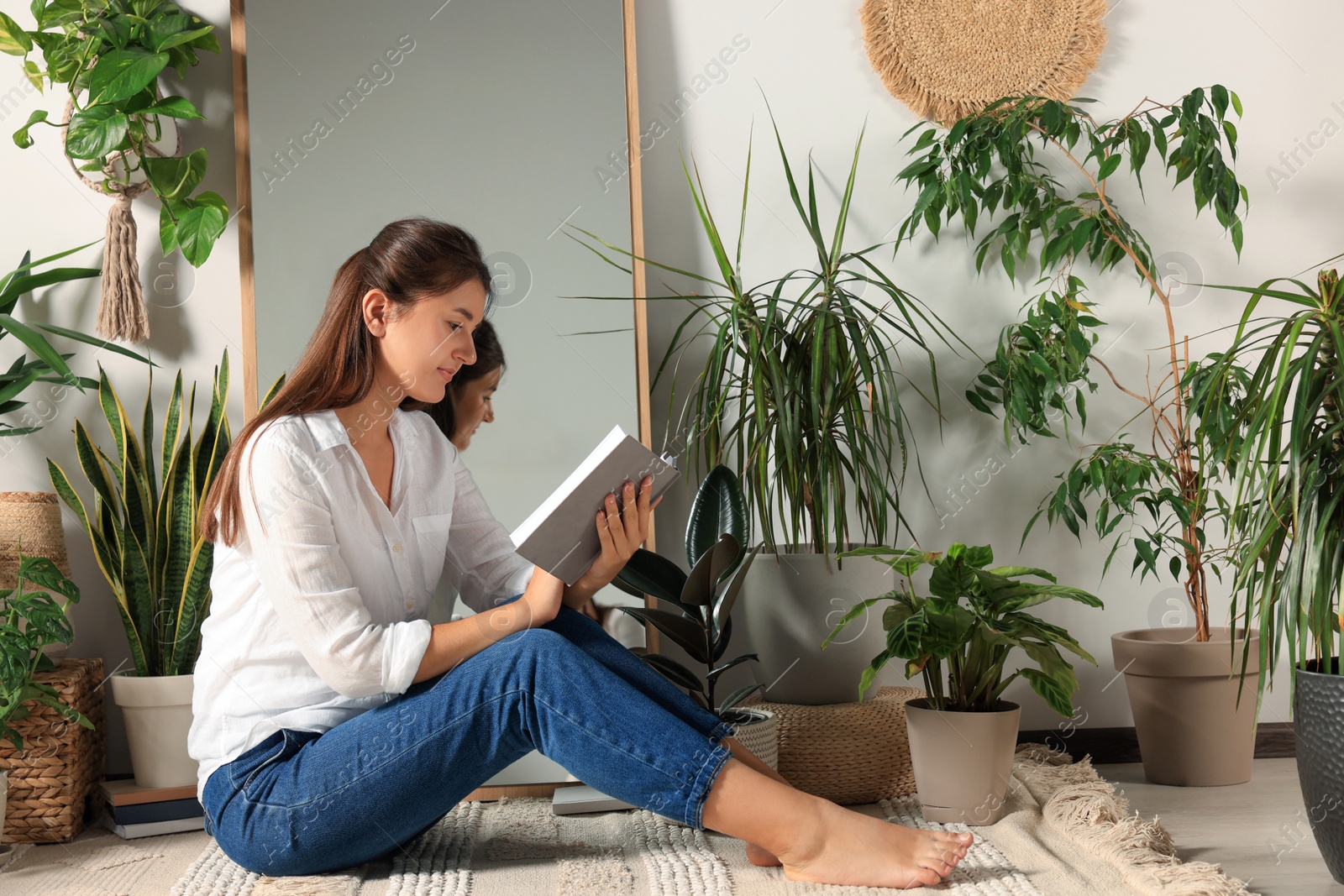 Photo of Young woman reading book near mirror and different houseplants on floor in room