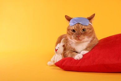 Photo of Cute ginger cat with sleep mask, crocheted bunny and red pillow on orange background, space for text