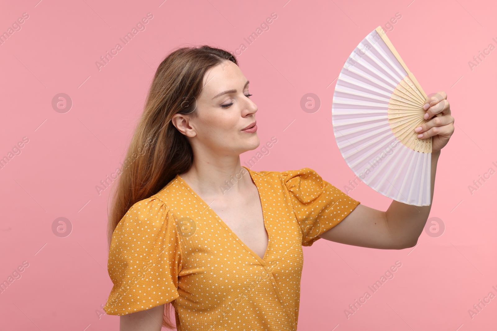 Photo of Beautiful woman waving yellow hand fan to cool herself on pink background