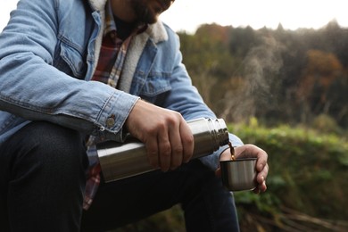 Man pouring hot drink from metallic thermos into cup lid in nature, closeup