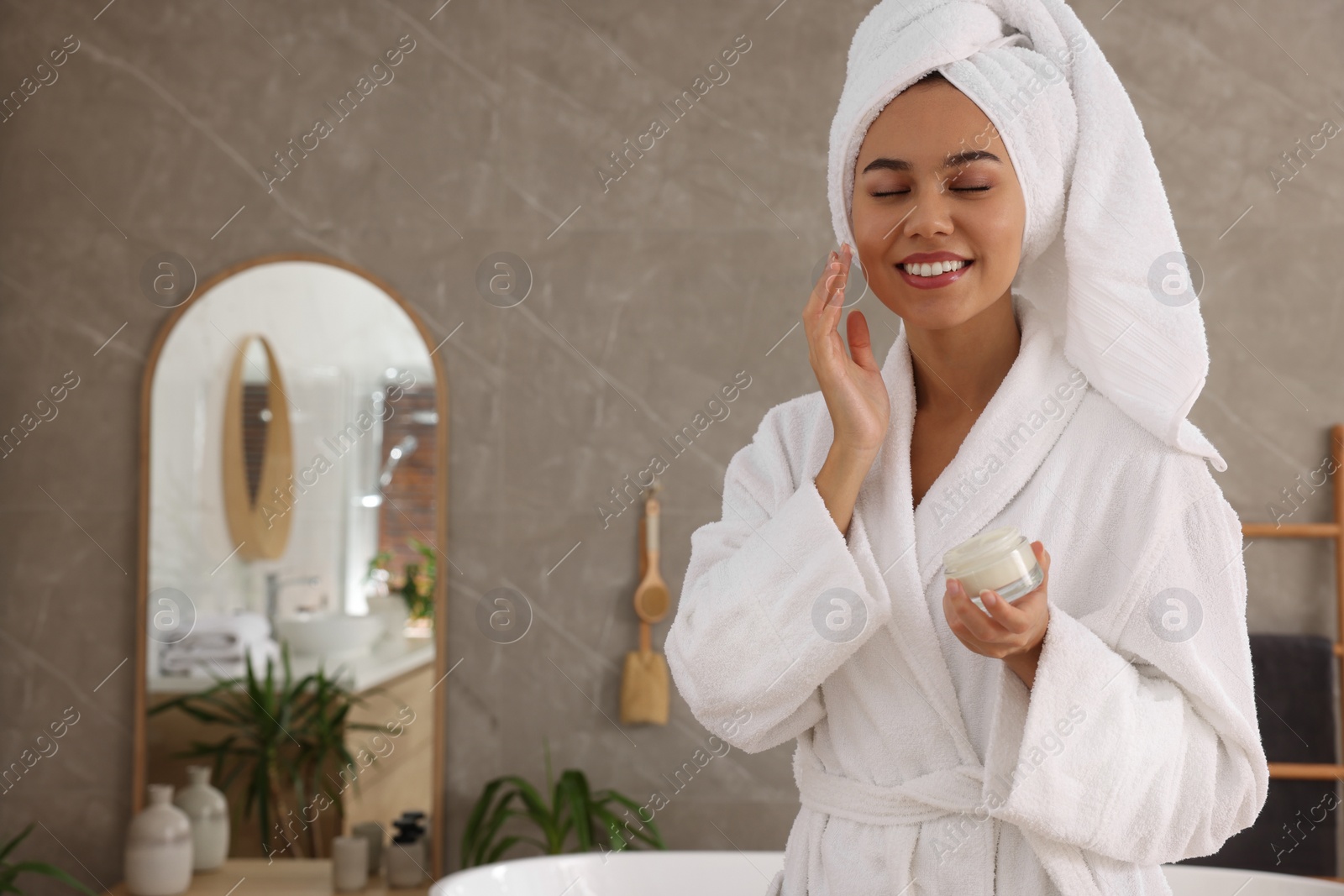 Photo of Beautiful African American woman applying cream onto face in bathroom, space for text