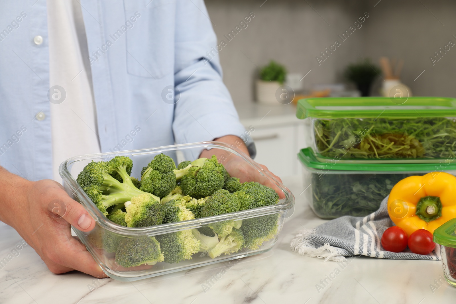 Photo of Man holding glass container with fresh broccoli at white marble table in kitchen, closeup. Food storage