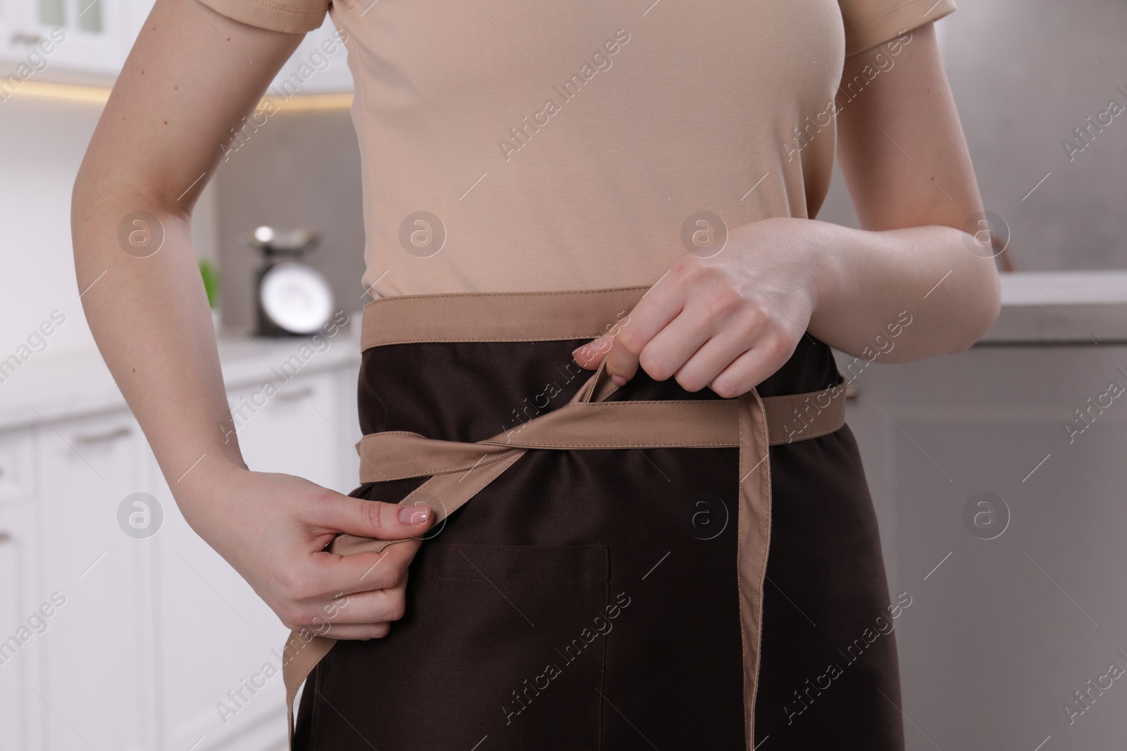 Photo of Woman putting on brown apron in kitchen , closeup