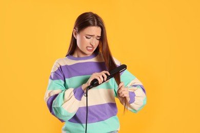 Stressed young woman with flattening iron on yellow background. Hair damage