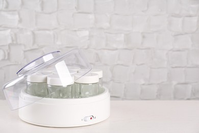 Modern yogurt maker with empty jars on white wooden table, space for text