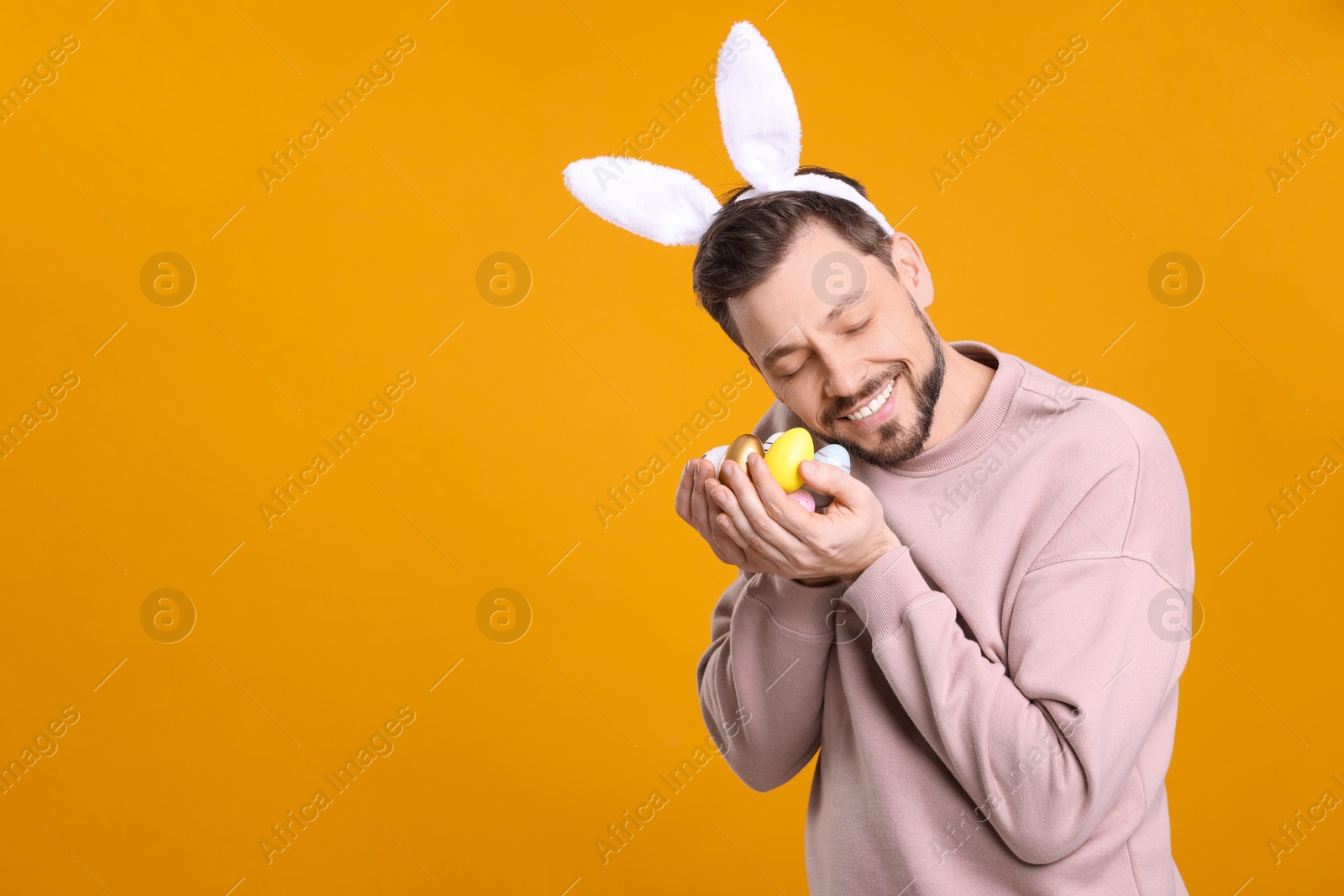 Photo of Happy man in bunny ears headband holding painted Easter eggs on orange background. Space for text