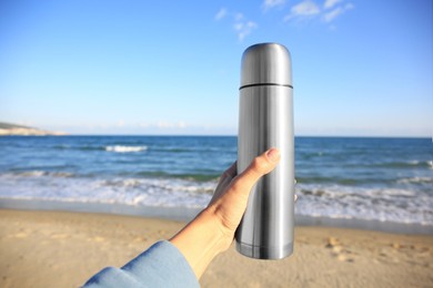 Woman holding metallic thermos with hot drink on beach near sea, closeup. Space for text