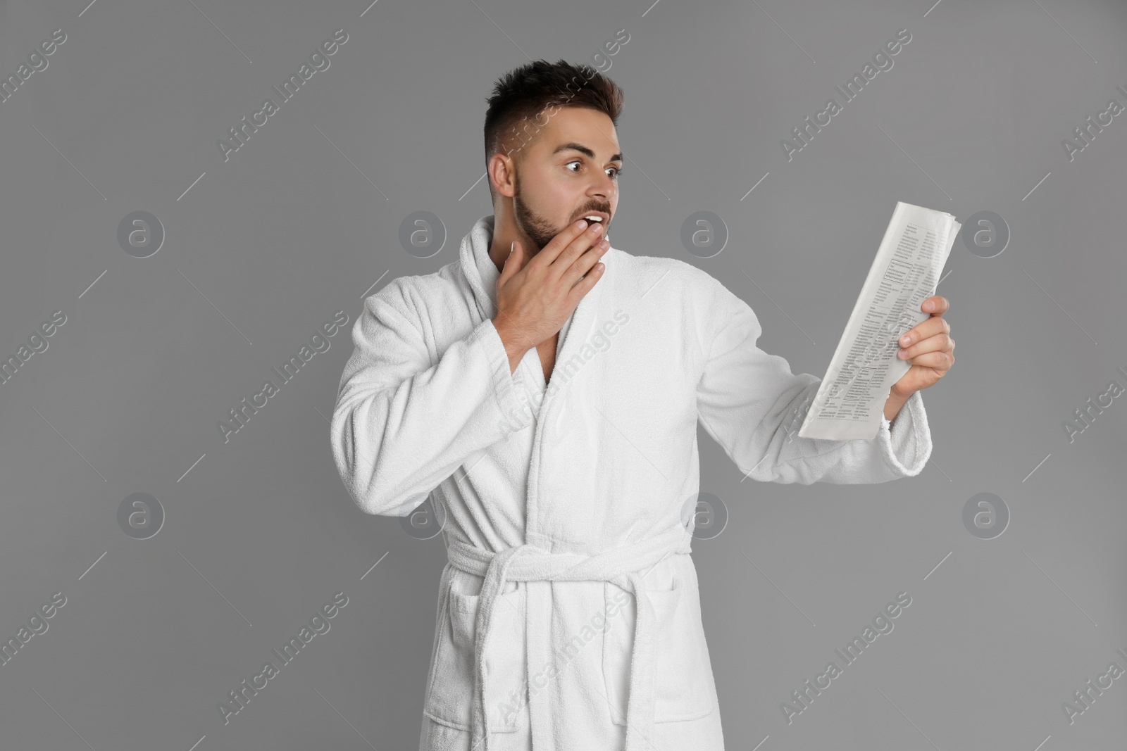 Photo of Young man in bathrobe reading newspaper on grey background