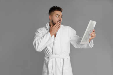 Young man in bathrobe reading newspaper on grey background