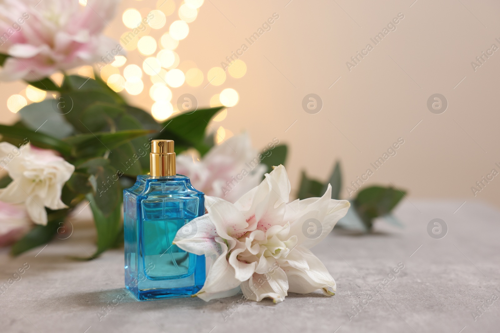 Photo of Bottle of perfume and beautiful lily flowers on table against beige background with blurred lights, closeup. Space for text