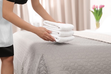 Young maid putting stack of fresh towels on bed in hotel room, closeup