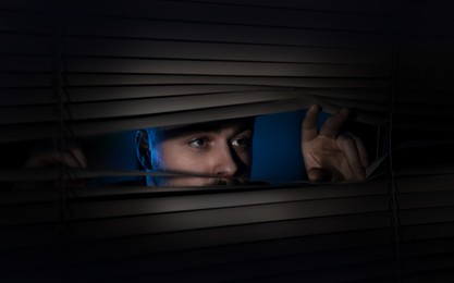 Image of Worried man looking through window blinds into darkness. Paranoia concept