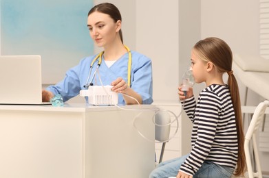 Medical assistant sitting near sick little girl while she using nebulizer for inhalation in hospital