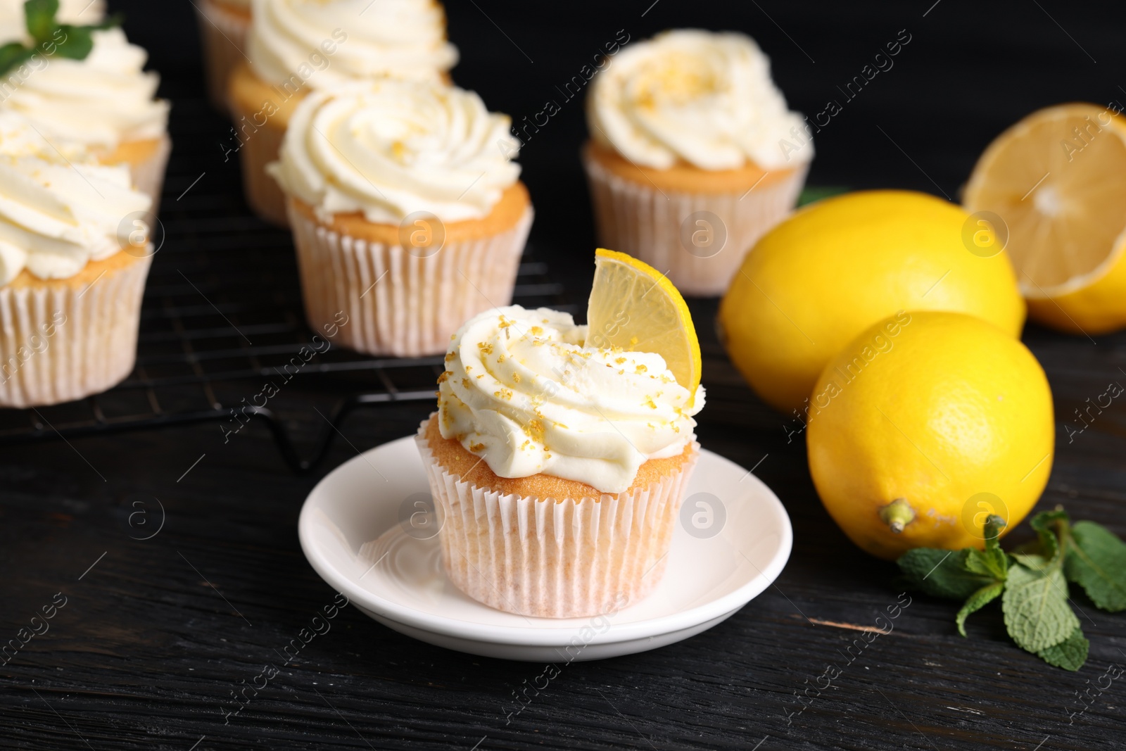 Photo of Delicious cupcakes with white cream, lemon zest and lemons on black wooden table, closeup