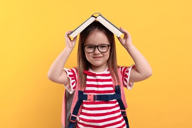 Cute little girl in glasses with open book and backpack against orange background