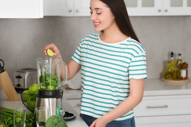 Beautiful young woman adding lime into blender with ingredients for smoothie in kitchen