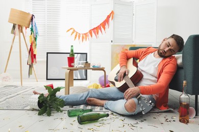 Photo of Young man with guitar sleeping near sofa in messy room after party