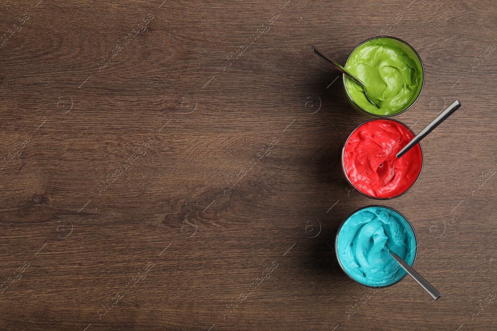 Photo of Glasses of different cream with food coloring on wooden table, flat lay. Space for text