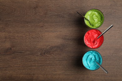 Glasses of different cream with food coloring on wooden table, flat lay. Space for text