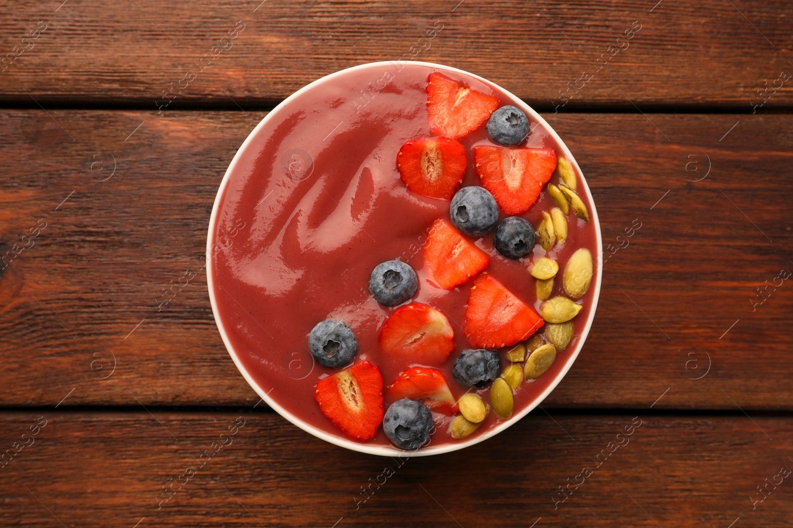 Photo of Bowl of delicious smoothie with fresh blueberries, strawberries and pumpkin seeds on wooden table, top view