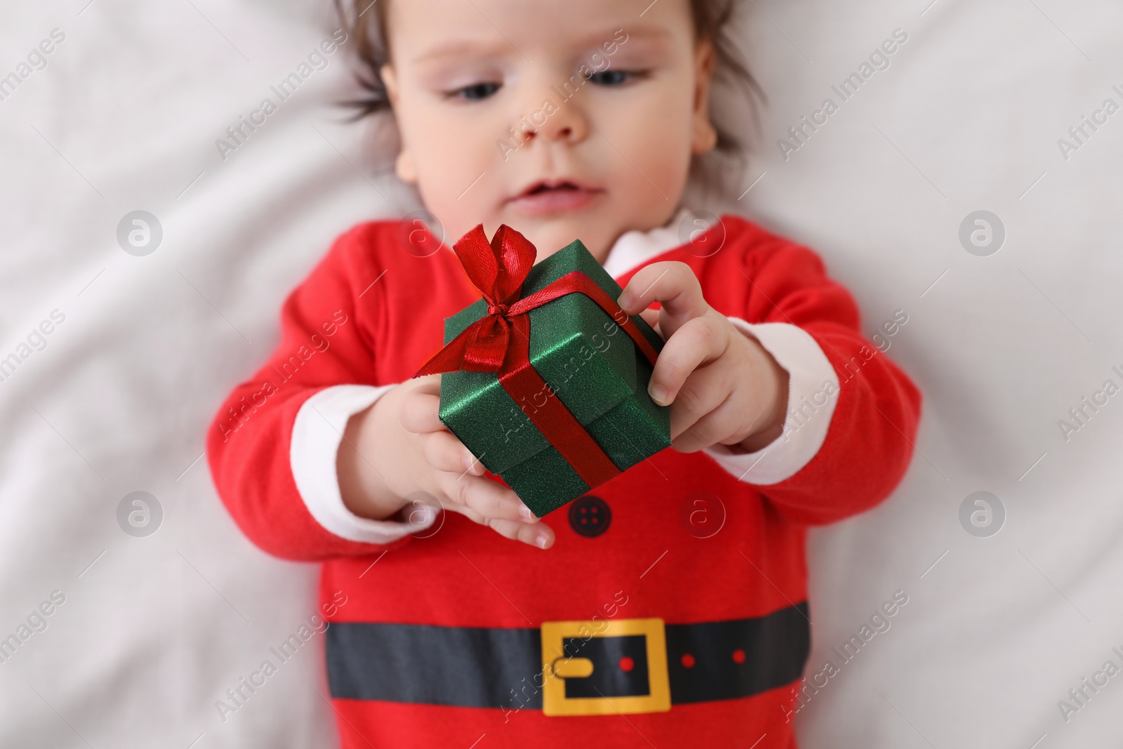 Photo of Cute baby wearing festive Christmas costume with gift box on white bedsheet, top view