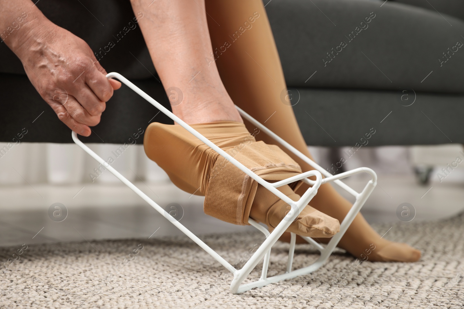 Photo of Woman putting on compression tights with stocking donner in living room, closeup. Prevention of varicose veins